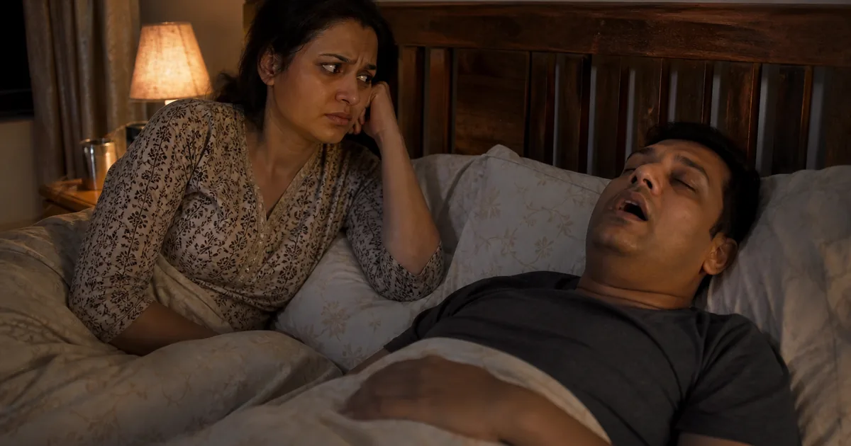 Indian couple in a dim bedroom, partner awake and concerned while the other sleeps on their back