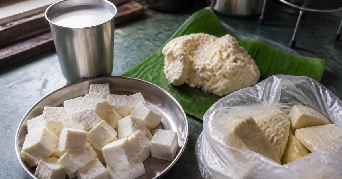 Fresh paneer cubes, a steel glass of milk, and khoya on an Indian kitchen counter beside a wet-market bag of pale cubes